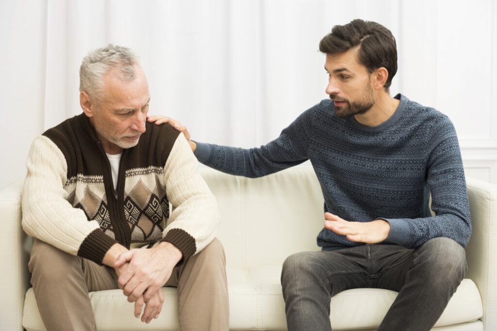 Ein Mann sitzt neben einem älteren Mann auf dem Sofa und legt ihm die Hand auf die Schulter